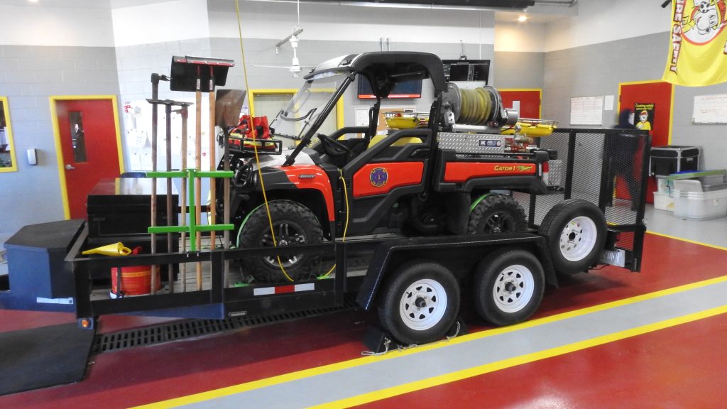 A fire rescue utility vehicle equipped with firefighting tools and water hoses is secured on a flatbed trailer inside a garage. The trailer is attached to a towing hitch, and various fire equipment is visible in the background.