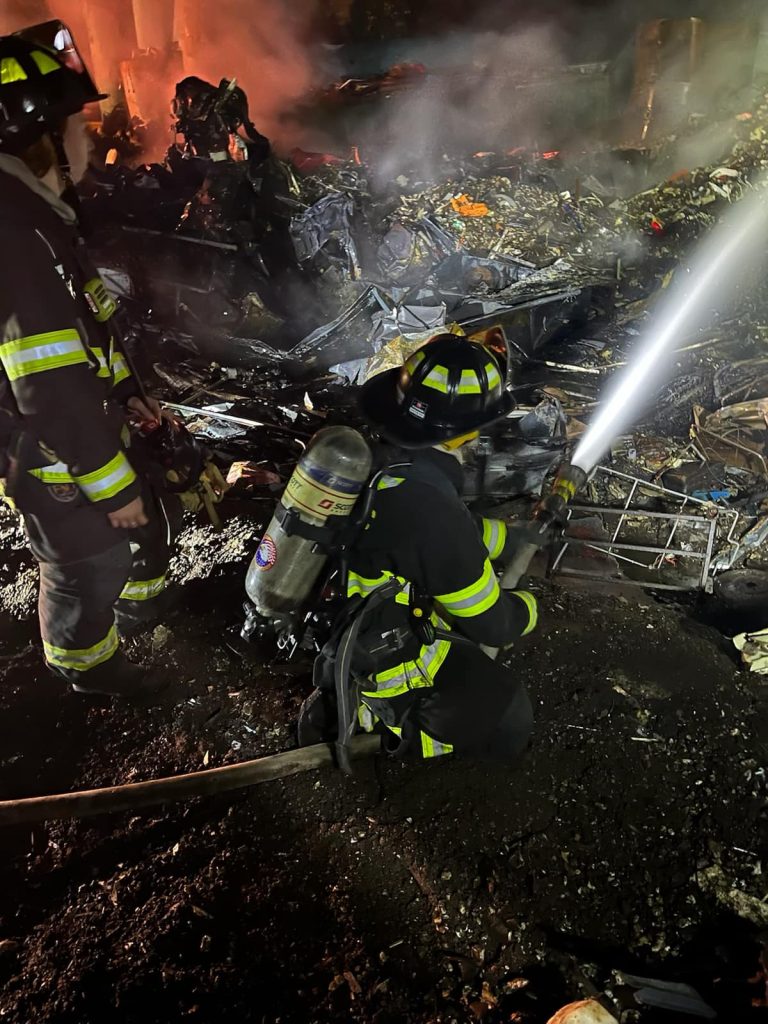 Two firefighters in full gear work to extinguish a blaze amidst smoking debris and rubble. One kneels, directing a hose at the fire, while the other stands nearby. The scene is dark, illuminated by the fire's glow.