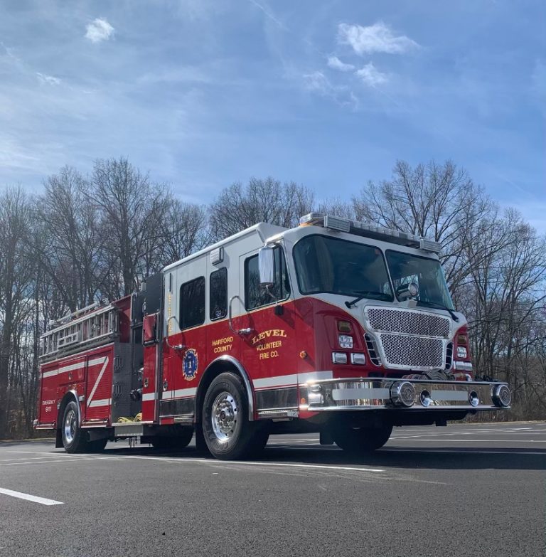 A red fire truck with silver accents parked on a paved surface, surrounded by leafless trees and under a clear blue sky. The truck has "Harford County" and "Level Volunteer Fire Co." written on its side.