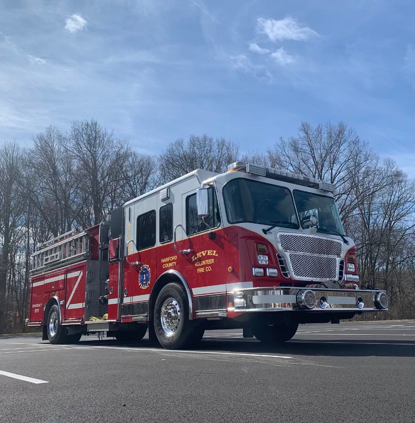 A red fire truck with silver accents parked on a paved surface, surrounded by leafless trees and under a clear blue sky. The truck has "Harford County" and "Level Volunteer Fire Co." written on its side.
