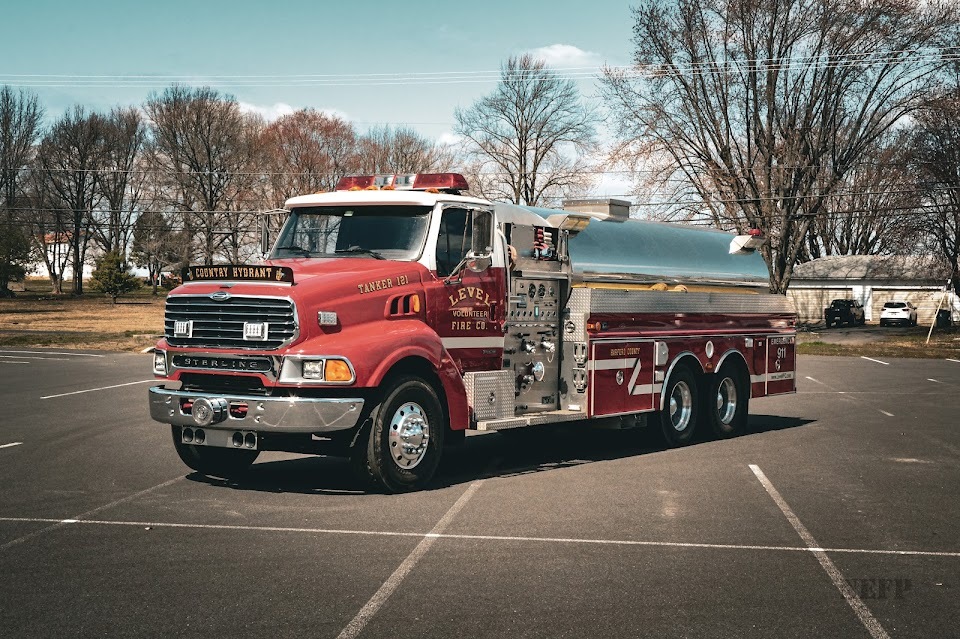 A red fire tanker truck parked in an empty lot on a clear day, surrounded by leafless trees. The truck is labeled with the number 111 and has metal tanks and equipment. The background includes power lines and a white building.
