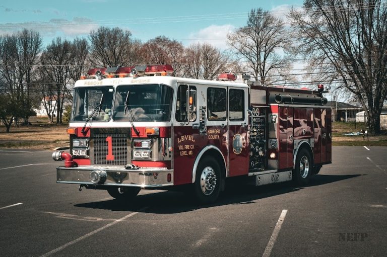 A red fire truck with a number 1 on its front is parked in an open area. The vehicle is equipped with lights and hoses, and the surrounding area includes leafless trees and a grassy field.