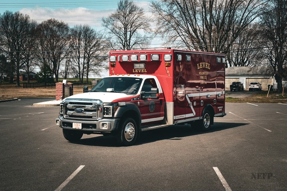 A red and white ambulance marked "Level Volunteer Fire Department" is parked in an empty lot. Trees without leaves and a small building are visible in the background under a partly cloudy sky.