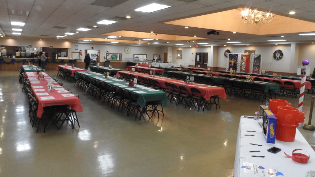 A banquet hall with long tables covered in red and green tablecloths, set with plates and cutlery. The room is decorated for a festive event, with a chandelier overhead and various decorations visible in the background.