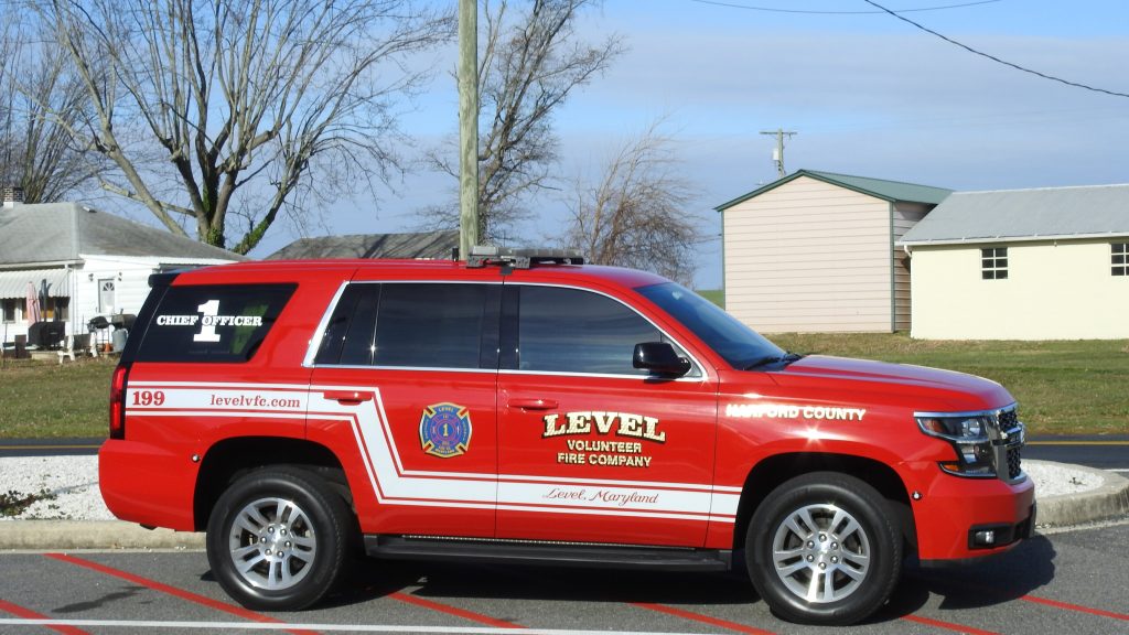 A red SUV marked "Level Volunteer Fire Company" with "Chief Officer" on the rear window is parked on a street. The vehicle has a fire department emblem and website. Buildings and trees are in the background.