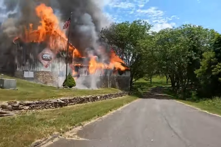 A wooden building engulfed in flames with thick smoke is situated beside a road lined with trees and a stone wall. An American flag is flying in front of the burning structure. Clear blue sky is visible in the background.