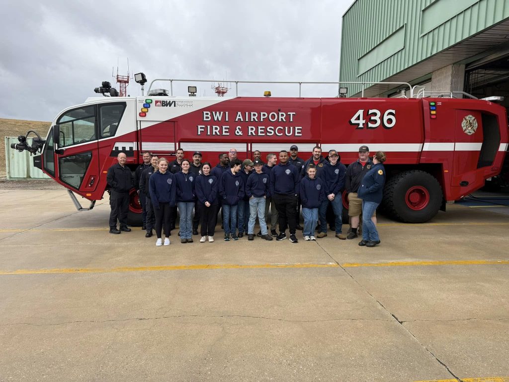 A group of people in matching blue uniforms stand in front of a large red BWI Airport Fire & Rescue vehicle marked with the number 436. The setting appears to be outside a building with a large garage door.