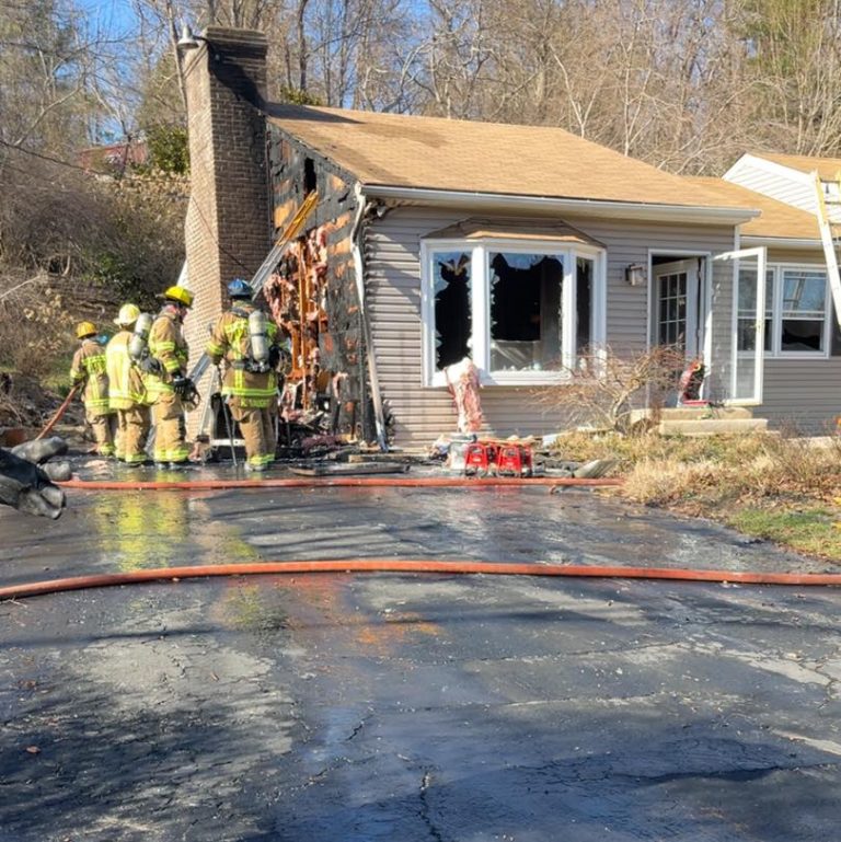 Firefighters in yellow gear inspect a small house with visible fire damage on its side and front. The siding is charred, and a ladder leans against the house. A fire hose and wet driveway are in the foreground, with trees in the background.