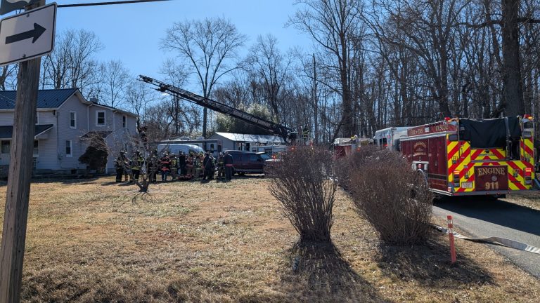 A group of firefighters gathers near a house with a ladder truck and fire engine parked on the side. The scene is on a grassy area with leafless trees in the background under a clear sky.